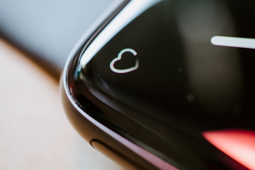 A close-up of the edge of a black smartwatch screen featuring a heart symbol, suggesting a focus on heart rate monitoring. The screen's curved edge reflects light, creating a sleek and modern appearance.