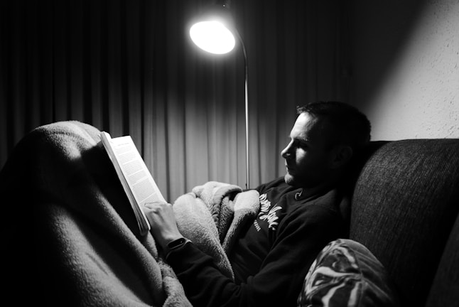 A person relaxing on a sofa reading an ebook on a digital device with a warm lamp glow.