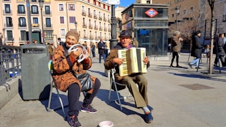 Two elderly street musicians are performing in an urban setting. One is playing an accordion while the other is holding a tambourine and smiling. They are seated on foldable chairs. The weather appears to be sunny, and people are walking by in the background. There's a trash can nearby and a subway entrance visible, with a sign indicating it's a metro station.