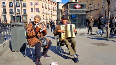 Two elderly street musicians are performing in an urban setting. One is playing an accordion while the other is holding a tambourine and smiling. They are seated on foldable chairs. The weather appears to be sunny, and people are walking by in the background. There's a trash can nearby and a subway entrance visible, with a sign indicating it's a metro station.