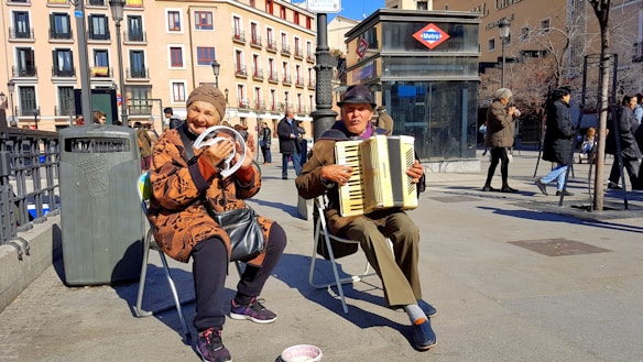 Two elderly street musicians are performing in an urban setting. One is playing an accordion while the other is holding a tambourine and smiling. They are seated on foldable chairs. The weather appears to be sunny, and people are walking by in the background. There's a trash can nearby and a subway entrance visible, with a sign indicating it's a metro station.