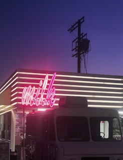 A neon pink sign spelling 'milk' is prominently displayed on a building with horizontal lights. The sky in the background transitions to a deep blue, and a utility pole with wires is visible. An ice cream truck or similar vehicle is parked in front, partially obscuring the storefront.