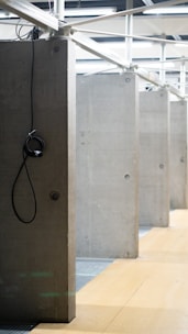 A series of concrete partitions are aligned in a row. Each partition has a hook hanging by a black cable. The flooring consists of wooden panels, and the ceiling features exposed structural elements with overhead lighting.