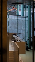 A modern interior space featuring large glass windows that offer a view of construction equipment outside. Inside, there is a sleek, white staircase leading to a sitting area with a single chair and several stacks of books arranged neatly on white tables. Potted plants are strategically placed to add a touch of greenery.