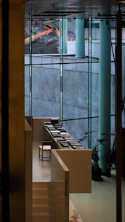 A modern interior space featuring large glass windows that offer a view of construction equipment outside. Inside, there is a sleek, white staircase leading to a sitting area with a single chair and several stacks of books arranged neatly on white tables. Potted plants are strategically placed to add a touch of greenery.