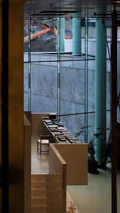 A modern interior space featuring large glass windows that offer a view of construction equipment outside. Inside, there is a sleek, white staircase leading to a sitting area with a single chair and several stacks of books arranged neatly on white tables. Potted plants are strategically placed to add a touch of greenery.