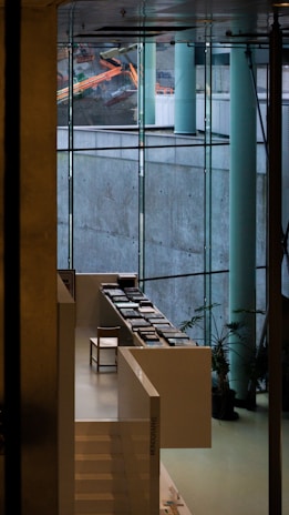 A modern interior space featuring large glass windows that offer a view of construction equipment outside. Inside, there is a sleek, white staircase leading to a sitting area with a single chair and several stacks of books arranged neatly on white tables. Potted plants are strategically placed to add a touch of greenery.