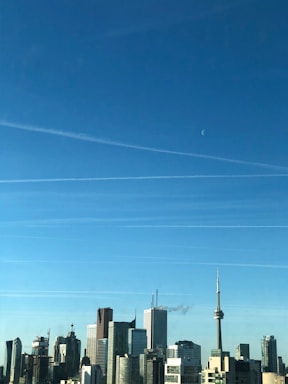 A city skyline with modern skyscrapers against a clear blue sky. The CN Tower is prominently visible among the buildings. Thin white contrails are spread across the sky, and a faint crescent moon is visible.
