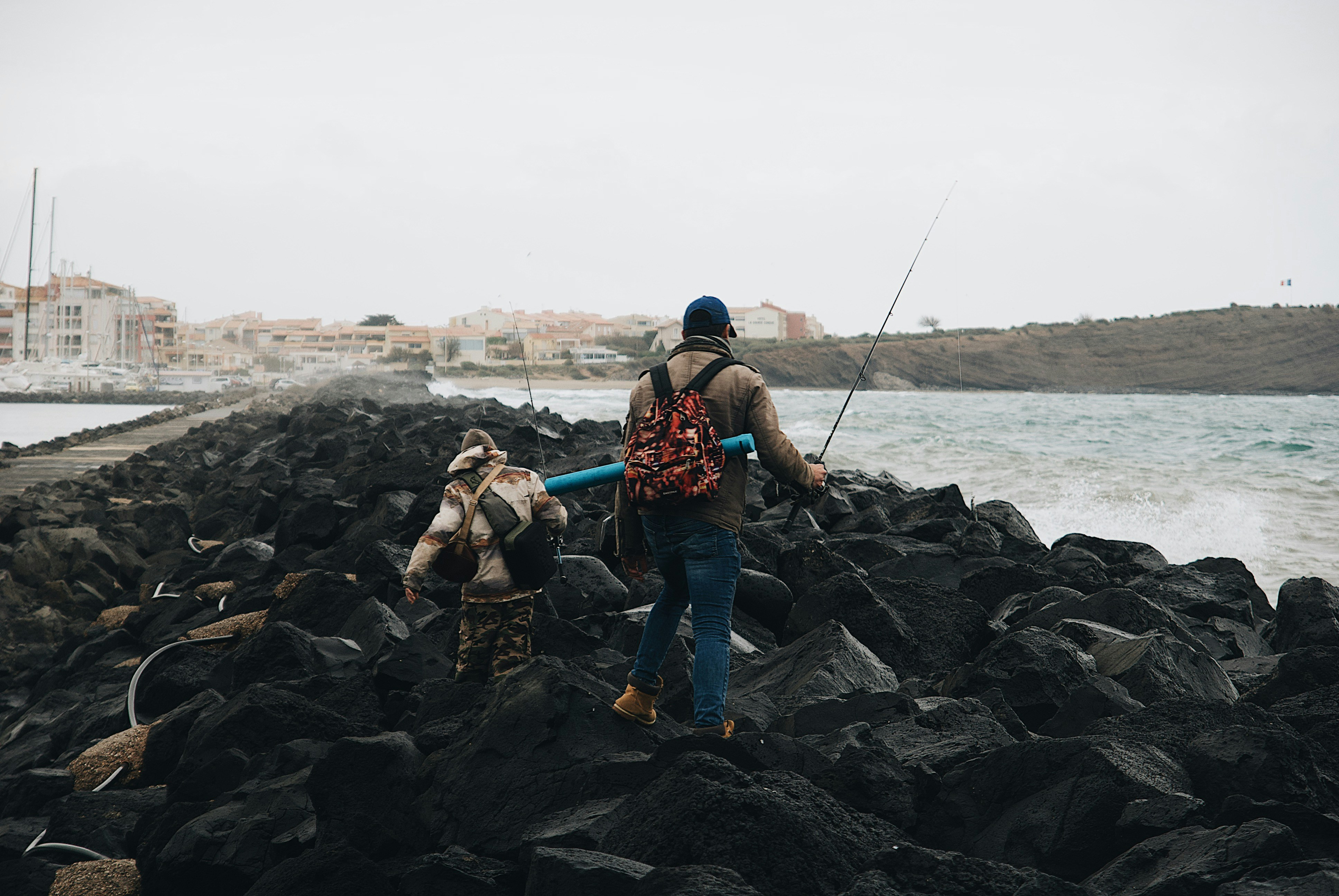 Two anglers with backpacks and rods climbing over black boulders on a harbor jetty, one carrying a blue rod tube, waves breaking on the right