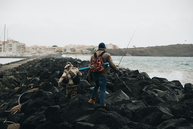 Two anglers with backpacks and rods climbing over black boulders on a harbor jetty, one carrying a blue rod tube, waves breaking on the right