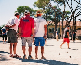 Three men stand with their backs to the camera, holding metal balls in their hands, likely playing a game of pétanque, outdoors on a sandy court surrounded by trees and a building in the background. Other people are visible in the distance, participating in or observing the activities.