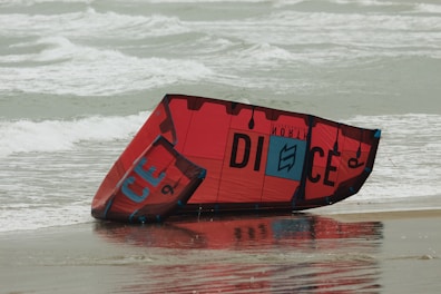 A red and black kiteboarding kite rests on a sandy beach with ocean waves in the background. The kite is partially wet, reflecting some light from the overcast sky.