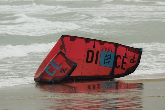A red and black kiteboarding kite rests on a sandy beach with ocean waves in the background. The kite is partially wet, reflecting some light from the overcast sky.