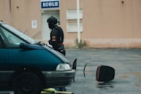A person dressed in dark protective gear and helmet is standing next to a teal vehicle in a wet parking lot. In the background, a beige building features a sign marked 'Ecole.' Nearby, a large object resembling a circular traffic paddle is lying on the ground.