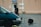 A person dressed in dark protective gear and helmet is standing next to a teal vehicle in a wet parking lot. In the background, a beige building features a sign marked 'Ecole.' Nearby, a large object resembling a circular traffic paddle is lying on the ground.