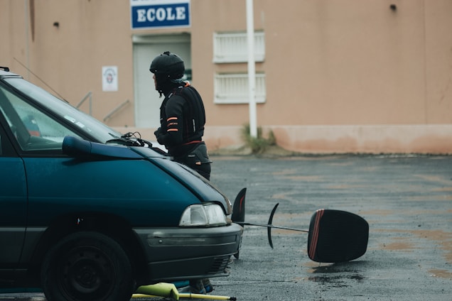 A person dressed in dark protective gear and helmet is standing next to a teal vehicle in a wet parking lot. In the background, a beige building features a sign marked 'Ecole.' Nearby, a large object resembling a circular traffic paddle is lying on the ground.