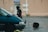 A person dressed in dark protective gear and helmet is standing next to a teal vehicle in a wet parking lot. In the background, a beige building features a sign marked 'Ecole.' Nearby, a large object resembling a circular traffic paddle is lying on the ground.