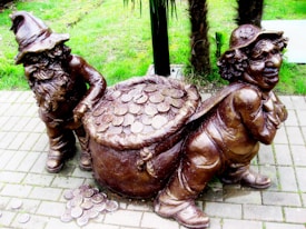 Two bronze statues of whimsical gnome-like figures are positioned on a paved surface next to a large, overflowing sack of coins. One figure wears a pointed hat and leans forward with one hand on the sack, while the other, with a rounded hat, appears to hold onto the sack. The ground is scattered with additional coins, and the background features grassy foliage.