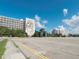 A spacious plaza with large buildings featuring prominent murals. The buildings are surrounded by lush greenery, and a few people can be seen walking around the open concrete area under a clear blue sky.