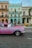 An old-fashioned pink car is parked on a street in front of pastel-colored colonial buildings, including a blue and beige facade. A yellow taxi is visible in the background along with several people walking on the sidewalk.