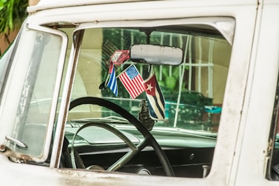 The interior of a vintage car is visible through the side window, featuring a steering wheel and three hanging flags from the rearview mirror including the American and Cuban flags. The car appears to have an aged and rustic look, with signs of wear and slightly faded colors.