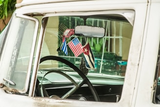The interior of a vintage car is visible through the side window, featuring a steering wheel and three hanging flags from the rearview mirror including the American and Cuban flags. The car appears to have an aged and rustic look, with signs of wear and slightly faded colors.