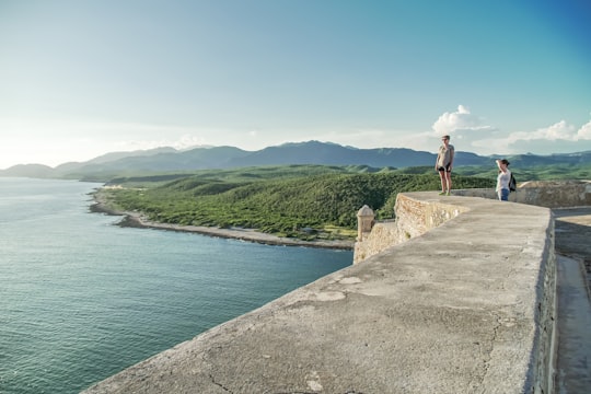 man in white t-shirt and brown shorts standing on concrete dock near body of water in Castillo de San Pedro de la Roca Cuba