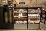 A gourmet grocery display with shelves stocked with various food items, including sauces, pastas, snack packages, and bottled drinks. Several shoppers can be seen browsing and carrying baskets nearby.