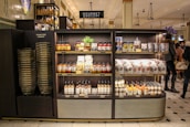 A gourmet grocery display with shelves stocked with various food items, including sauces, pastas, snack packages, and bottled drinks. Several shoppers can be seen browsing and carrying baskets nearby.