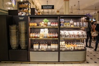 A gourmet grocery display with shelves stocked with various food items, including sauces, pastas, snack packages, and bottled drinks. Several shoppers can be seen browsing and carrying baskets nearby.