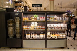 A gourmet grocery display with shelves stocked with various food items, including sauces, pastas, snack packages, and bottled drinks. Several shoppers can be seen browsing and carrying baskets nearby.