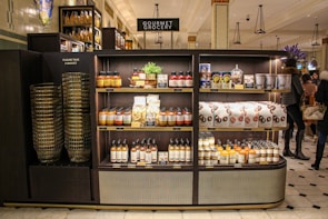 A gourmet grocery display with shelves stocked with various food items, including sauces, pastas, snack packages, and bottled drinks. Several shoppers can be seen browsing and carrying baskets nearby.