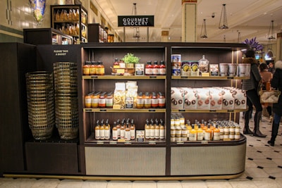 A gourmet grocery display with shelves stocked with various food items, including sauces, pastas, snack packages, and bottled drinks. Several shoppers can be seen browsing and carrying baskets nearby.