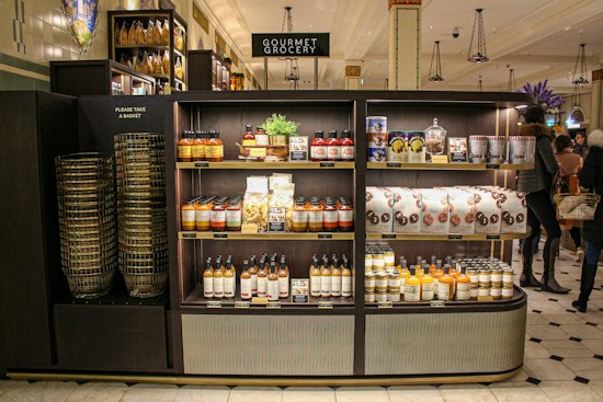 A gourmet grocery display with shelves stocked with various food items, including sauces, pastas, snack packages, and bottled drinks. Several shoppers can be seen browsing and carrying baskets nearby.