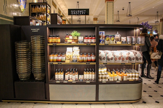A gourmet grocery display with shelves stocked with various food items, including sauces, pastas, snack packages, and bottled drinks. Several shoppers can be seen browsing and carrying baskets nearby.
