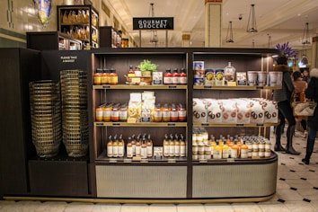 A gourmet grocery display with shelves stocked with various food items, including sauces, pastas, snack packages, and bottled drinks. Several shoppers can be seen browsing and carrying baskets nearby.