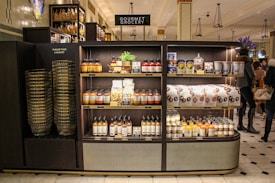 A gourmet grocery display with shelves stocked with various food items, including sauces, pastas, snack packages, and bottled drinks. Several shoppers can be seen browsing and carrying baskets nearby.