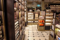 A well-organized interior of a luxury food store featuring shelves lined with jars and boxes. The store includes sections labeled 'The Bakery' and 'Biscuits' with neatly stacked products. A distinctive Harrods shopping bag is on display. The floor has a checkered tile pattern, and the lighting casts a warm, inviting glow.
