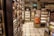 A well-organized interior of a luxury food store featuring shelves lined with jars and boxes. The store includes sections labeled 'The Bakery' and 'Biscuits' with neatly stacked products. A distinctive Harrods shopping bag is on display. The floor has a checkered tile pattern, and the lighting casts a warm, inviting glow.