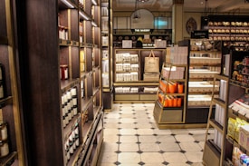 A well-organized interior of a luxury food store featuring shelves lined with jars and boxes. The store includes sections labeled 'The Bakery' and 'Biscuits' with neatly stacked products. A distinctive Harrods shopping bag is on display. The floor has a checkered tile pattern, and the lighting casts a warm, inviting glow.