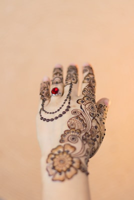 A hand decorated with intricate henna designs, featuring floral and paisley patterns. A red gemstone ring adorns one of the fingers, and a chain-like design adds elegance to the artwork. The background is a soft beige color, which highlights the detailed mehndi art.