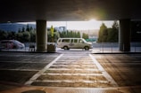 A van is parked under a covered area at an airport or transportation hub, with the sun setting in the background. The van is white with writing on its side. In the foreground, there is a crosswalk leading towards the van and a few shopping carts to the left. Tall trees and a modern building are visible in the background.