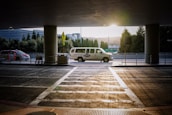A van is parked under a covered area at an airport or transportation hub, with the sun setting in the background. The van is white with writing on its side. In the foreground, there is a crosswalk leading towards the van and a few shopping carts to the left. Tall trees and a modern building are visible in the background.