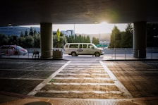 A van is parked under a covered area at an airport or transportation hub, with the sun setting in the background. The van is white with writing on its side. In the foreground, there is a crosswalk leading towards the van and a few shopping carts to the left. Tall trees and a modern building are visible in the background.