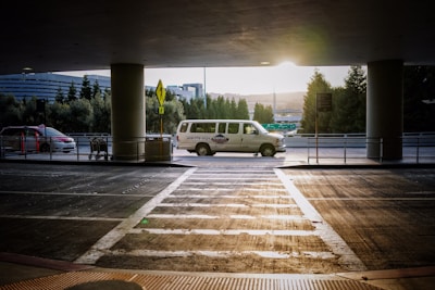 A van is parked under a covered area at an airport or transportation hub, with the sun setting in the background. The van is white with writing on its side. In the foreground, there is a crosswalk leading towards the van and a few shopping carts to the left. Tall trees and a modern building are visible in the background.