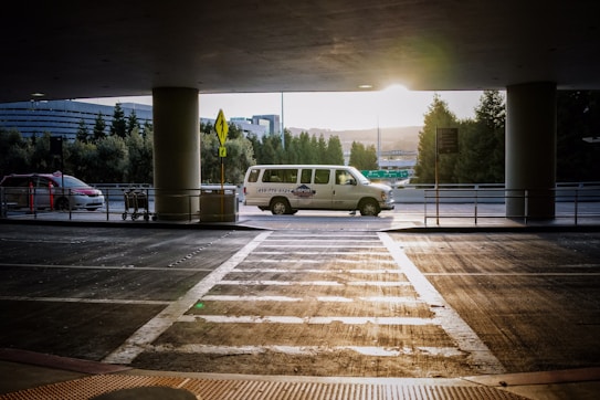A van is parked under a covered area at an airport or transportation hub, with the sun setting in the background. The van is white with writing on its side. In the foreground, there is a crosswalk leading towards the van and a few shopping carts to the left. Tall trees and a modern building are visible in the background.