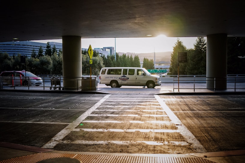 A van is parked under a covered area at an airport or transportation hub, with the sun setting in the background. The van is white with writing on its side. In the foreground, there is a crosswalk leading towards the van and a few shopping carts to the left. Tall trees and a modern building are visible in the background.