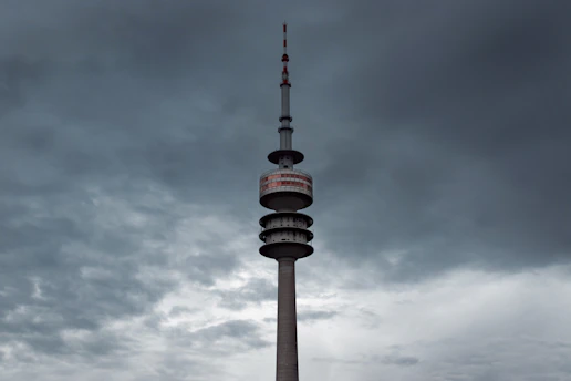 A tall telecommunications tower with multiple platforms and red and white markings rises against a backdrop of dark, overcast skies. The tower's structure is prominent and stands solitary amidst a sky filled with heavy clouds, creating a dramatic contrast.