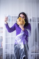 A person stands in a room holding a mesh bag of potatoes in front of their face and a medical mask connected to tubing in their other hand. The individual is wearing a purple shirt and is draped in clear plastic material. Bookshelves and other home items are visible in the background.