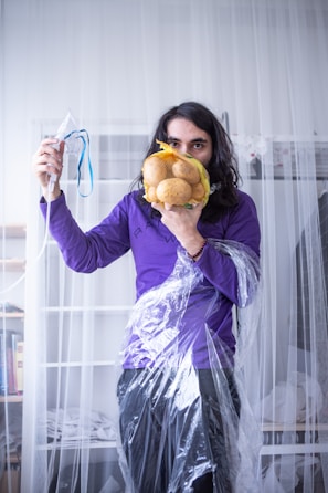 A person stands in a room holding a mesh bag of potatoes in front of their face and a medical mask connected to tubing in their other hand. The individual is wearing a purple shirt and is draped in clear plastic material. Bookshelves and other home items are visible in the background.
